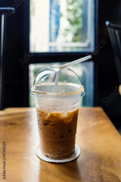 Obraz Iced Thai tea with straw in plastic cup on wooden table near window of cafe. Beverage.