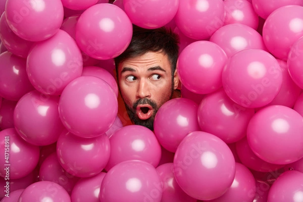 Fototapeta Surprised bearded European man sticks head through decorated background with inflated balloons prepares for holiday being shocked by unexpected relevation. People celebration emotions concept