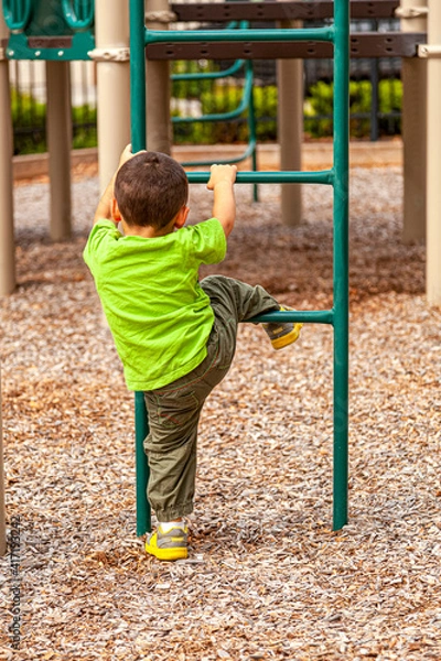 Fototapeta A white toddler boy is playing at a playground alone. He is holding the bars of a metal ladder and trying to pull himself up as he learns to climb. Surface is covered with wood chips for added safety