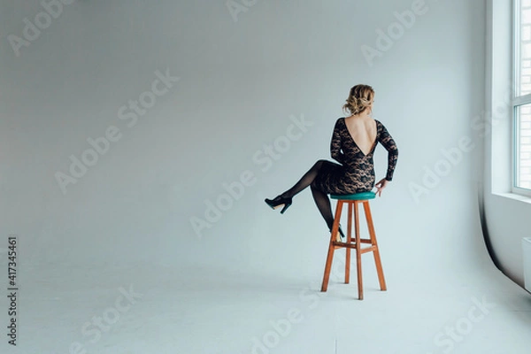 Obraz A young woman in a beautiful black guipure dress with an open back sits on a bar stool with her back to the viewer in an empty gray room
