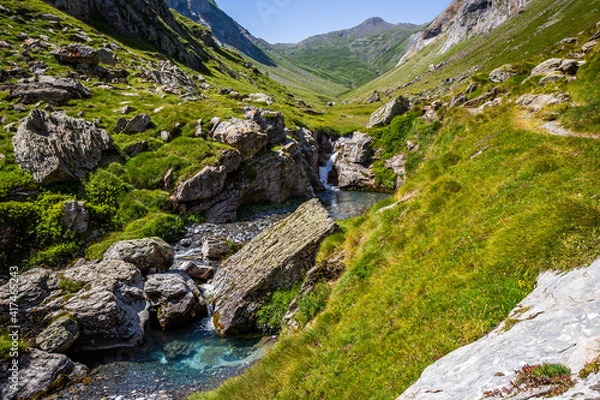 Obraz Beautiful turquoise blue waterfall surrounded by a green meadow with mountains in the background and a blue sky in springtime