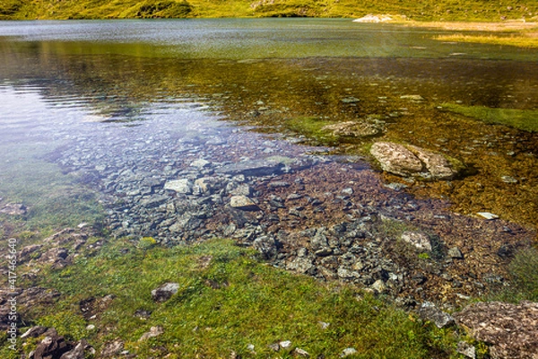 Obraz Crystal clear water of a lake surrounded by mountains, in a green meadow in summertime