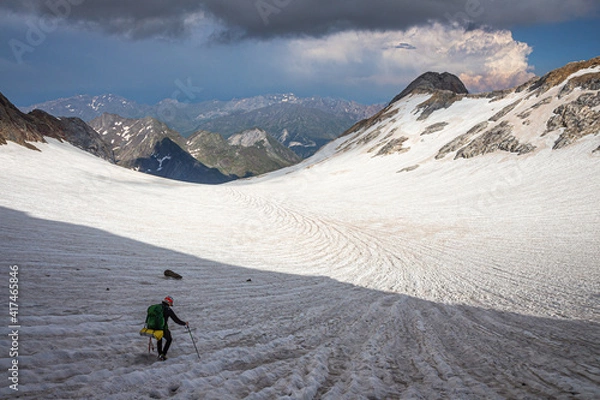 Obraz Climber crossing a glacier surrounded by mountains, with snow-capped mountains and a storm in the background
