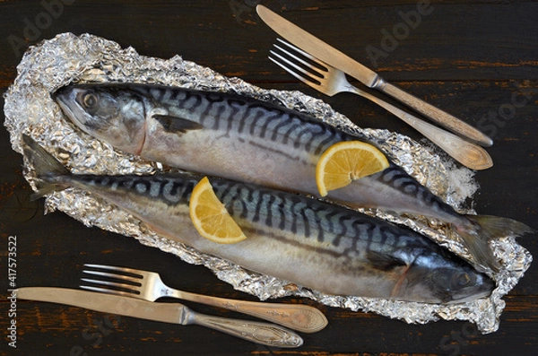 Fototapeta Two atlantic mackerel fishes ready for cooking decorated with spices, salt, lemon slices, knife and fork on the black wooden background. Top view