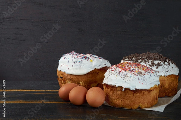 Fototapeta Traditional Easter bread or cake with white glaze and multicolored rainbow sprinkle decorated with eggs on the black wooden background. Close up view