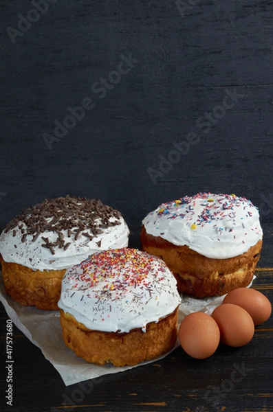 Fototapeta Traditional Ukrainian Easter bread or cake with white glaze and multicolored rainbow sprinkle decorated with eggs on the black wooden background. Side view with copy space