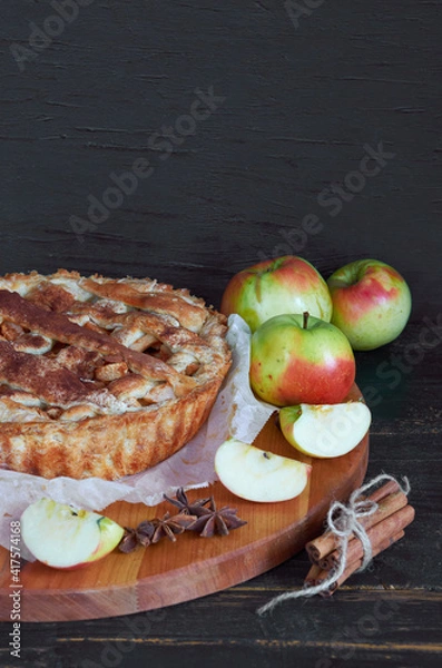 Fototapeta Traditional apple pie with spices on the brown wooden board. Apple pie decorated with sliced fresh apples and cinnamon sticks on the black background. Side view  with copy space