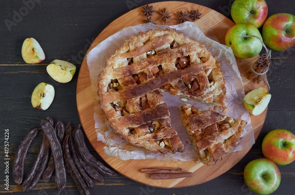 Fototapeta Traditional sliced apple pie with spices on the brown wooden board. Apple pie decorated with fresh apples, carob pods, anise stars and cinnamon sticks  on the black background. Top view 