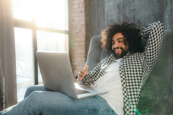 Fototapeta Smiling Egyptian in a plaid shirt sits in a chair working at the computer. Work remotely