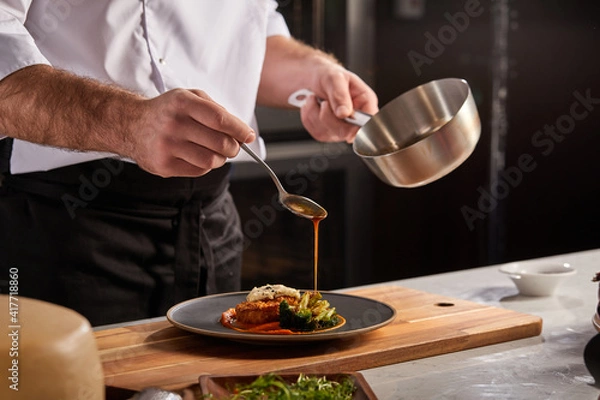 Fototapeta Cook in apron adding some sauce to dish. Cropped chef preparing food, meal, in kitchen, chef cooking, Chef decorating dish, closeup