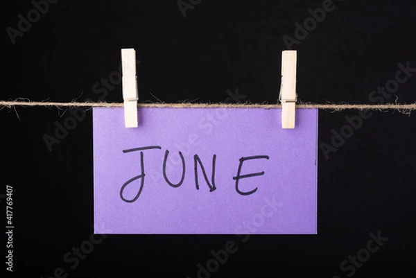 Fototapeta  June word written on a Purple color sticky note hanging with a wire on black background.