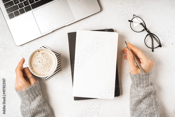 Fototapeta Top view woman hands with blank paper Notebook, leaf, laptop keyboard, cup of coffee, glasses and pen. Desktop mock up, Flat lay of white working table background with office equipment, mockup.