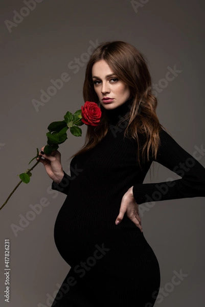 Obraz Pregnant girl in black tight dress. With a rose in my hand. Hand on the waist.  Grey background.