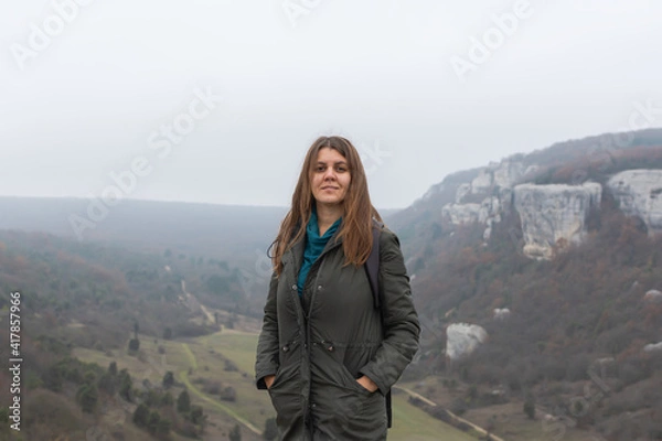 Fototapeta A young dark-haired woman on the background of an autumn landscape. Cloudy gray foggy view from the mountain. A happy smiling girl in a green coat. Natural background, concept of hiking, ecotourism