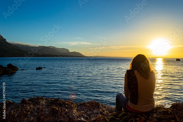 Fototapeta A young and beautiful girl dressed in casual clothes looking at the sunset at a beautiful beach in Artà Mallorca Balearic Islands Spain with a impressive view of the sea and the Tramuntana mountains