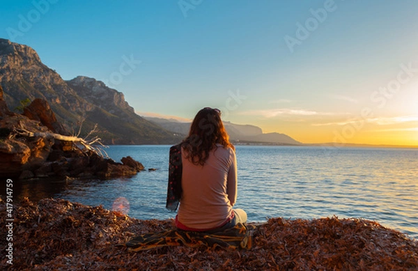 Obraz A young and beautiful girl dressed in casual clothes looking at the sunset at a beautiful beach in Artà Mallorca Balearic Islands Spain with a impressive view of the sea and the Tramuntana mountains