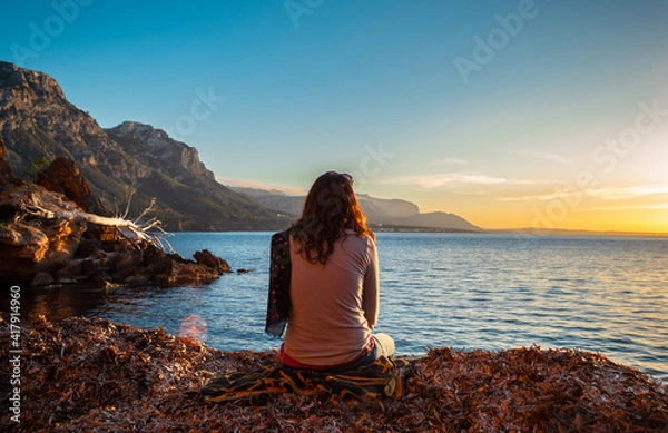 Fototapeta A young and beautiful girl dressed in casual clothes looking at the sunset at a beautiful beach in Artà Mallorca Balearic Islands Spain with a impressive view of the sea and the Tramuntana mountains