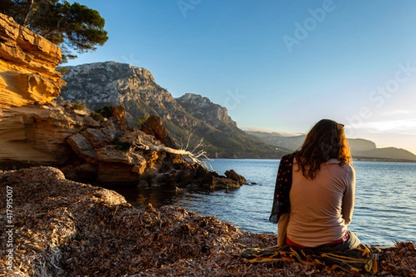 Obraz A young and beautiful girl dressed in casual clothes looking at the sunset at a beautiful beach in Artà Mallorca Balearic Islands Spain with a impressive view of the sea and the Tramuntana mountains