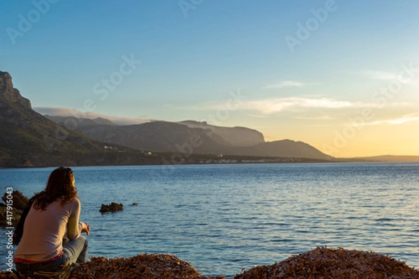 Fototapeta A young and beautiful girl dressed in casual clothes looking at the sunset at a beautiful beach in Artà Mallorca Balearic Islands Spain with a impressive view of the sea and the Tramuntana mountains