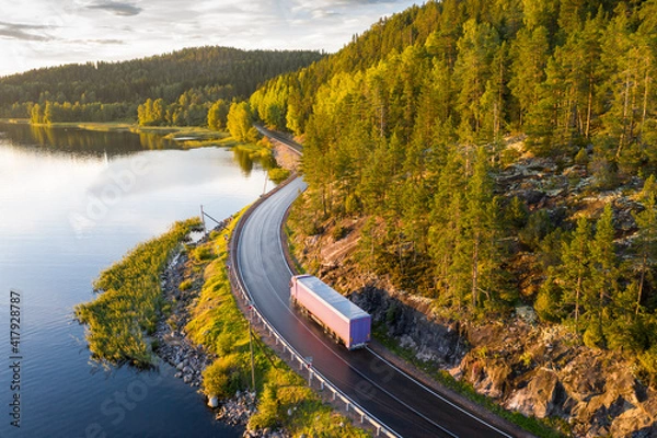 Fototapeta Aerial view of semi truck with cargo trailer on road curve at lake shore with green pine forest. Transportation background. Beautiful nature landscape at sunset in Republic of Karelia, Russia