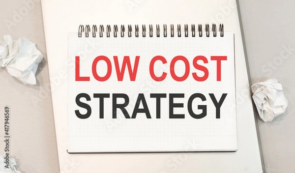 Fototapeta The word low cost strategy is written in a notebook that sits on a gray desktop along with a laptop.