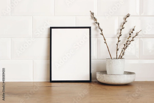 Obraz Spring still life. Blank black picture frame mockup on wooden table background. Easter composition with blooming goat willow, pussy willow or great sallow in ceramic vase. White tiles wall background.