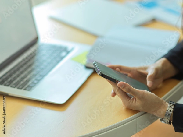 Obraz Businessman using smartphone in his hands on workspace with laptop and supplies