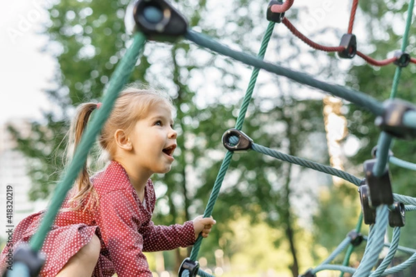 Fototapeta Children play and climb outdoors on a sunny summer day. Nice kid girl on a swing nest in a preschool sports complex.