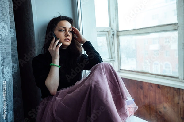 Fototapeta a depressed girl sits at the window and talking on the phone