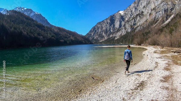 Fototapeta A woman walking along the shore of Leopoldsteiner lake in Austria. The lake is surrounded by high Alps. The shallow water is crystal clear, spring water has a calm surface. Early spring. Serenity