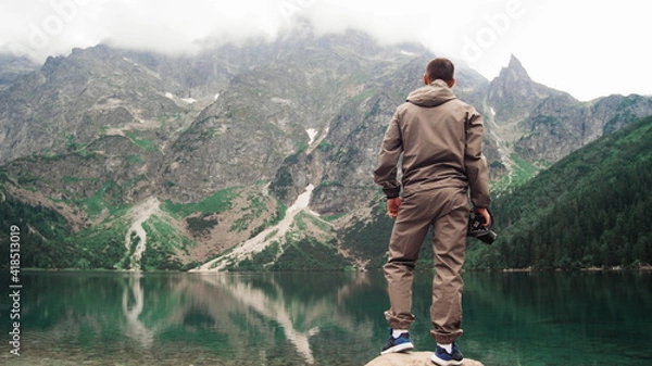 Obraz A man climbs a stone near the shore of a picturesque mountain lake with emerald water. Great views of nature. Travel and healthy lifestyle concept.