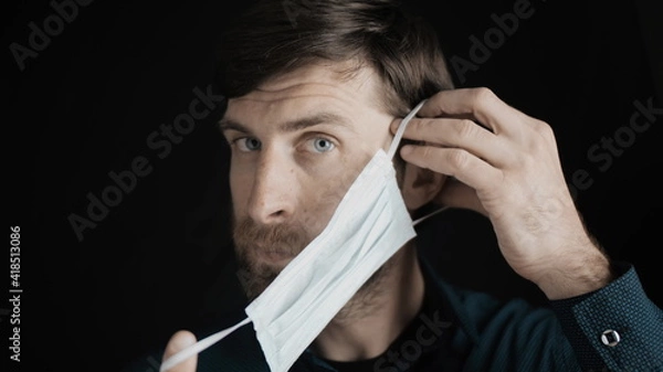 Obraz Attractive bearded man is putting on surgical mask on his face for corona virus covid-19 prevention. Infection protection, quarantine and epidemic concept. Slow motion close up front view 4k video.