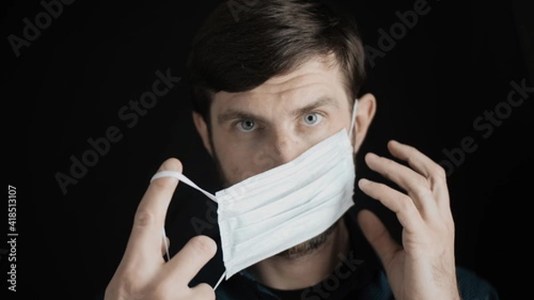 Obraz Attractive bearded man is putting on surgical mask on his face for corona virus covid-19 prevention. Infection protection, quarantine and epidemic concept. Slow motion close up front view 4k video.