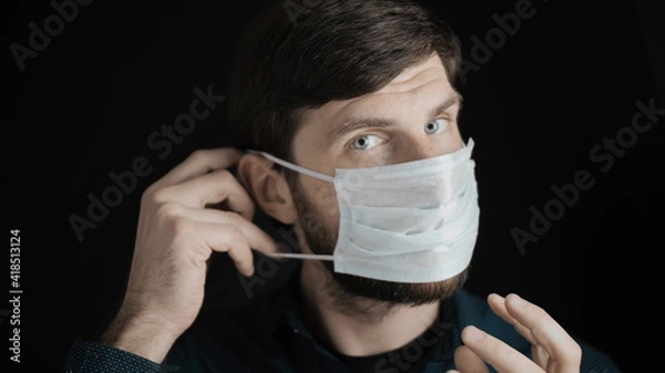 Obraz Attractive bearded man is putting on surgical mask on his face for corona virus covid-19 prevention. Infection protection, quarantine and epidemic concept. Slow motion close up front view 4k video.