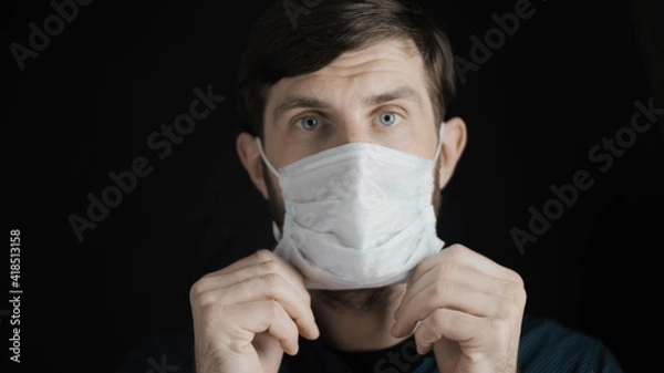 Obraz Attractive bearded man is putting on surgical mask on his face for corona virus covid-19 prevention. Infection protection, quarantine and epidemic concept. Slow motion close up front view 4k video.