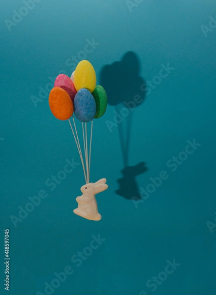 Fototapeta A white rabbit flies with balloons of colorful eggs. Blue background with shadow of bunny and easter eggs.