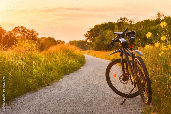 Fototapeta The mountain bike stands on a gravel bike path among green vegetation illuminated by the rays of the setting sun.