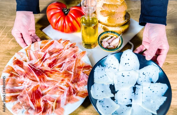 Fototapeta Typical Catalan or Spanish food. Bread with tomato, Iberian ham plate, goat cheese, olive oil and garlic. Top view of food products for dinner or tapas on a wooden background.