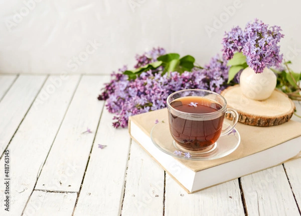 Fototapeta Romantic background with a cup of tea, lilac flowers and a book over a white wooden table. Leisure concept, spring breakfast