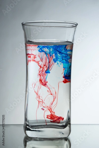 Fototapeta Single drops of light blue and red ink falling down into the glass full of water in the studio. Reflecting surface and gradient background