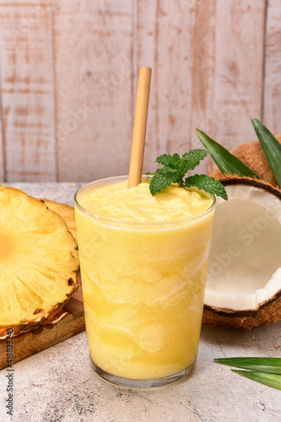 Obraz pineapple coconut smoothie with a bamboo straw and a mint leaf on top. on the side pineapple slices and two coconuts. stone surface and white wooden planks background. copy space for advertising.
