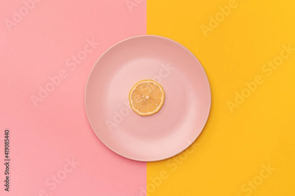 Fototapeta Slice of the fresh lemon on a pink plate. The background is divided vertically into pink and yellow. The lemon in the center of the frame. Minimalism