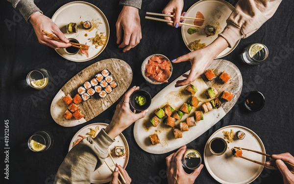 Fototapeta Family lockdown Japanese sushi dinner from delivery service at home. Flat-lay of table with salmon, crab, prawn, vegan rolls, wasabi, ginger and people eating together over dark background, top view