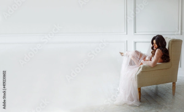 Obraz A bride in a white interior in a beautiful boudoir semi-transparent dress. She is sitting on the sofa, she has a beautiful wedding hairstyle and makeup