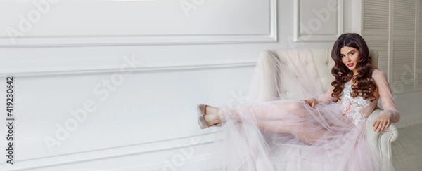 Obraz A bride in a white interior in a beautiful boudoir semi-transparent dress. She is sitting on the sofa, she has a beautiful wedding hairstyle and makeup