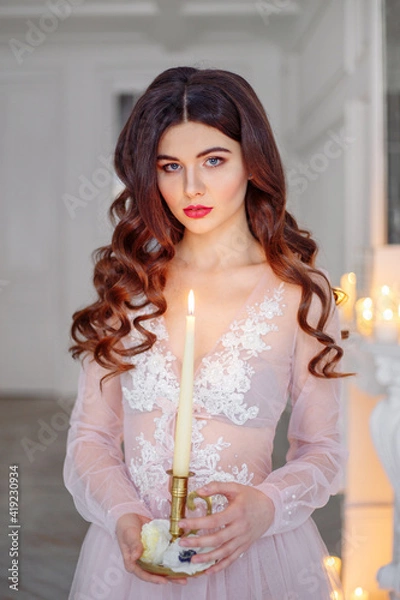 Obraz Portrait of a bride in a white interior in a beautiful boudoir semi-transparent dress. She holds a candle in her hand