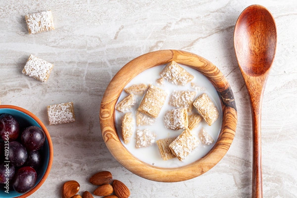 Fototapeta A simple breakfast concept with a bowl of frosted bite size wheat cereal in milk on marble kitchen countertop. There are grapes and nuts as appetizers. A healty snack with vitamins and fiber.