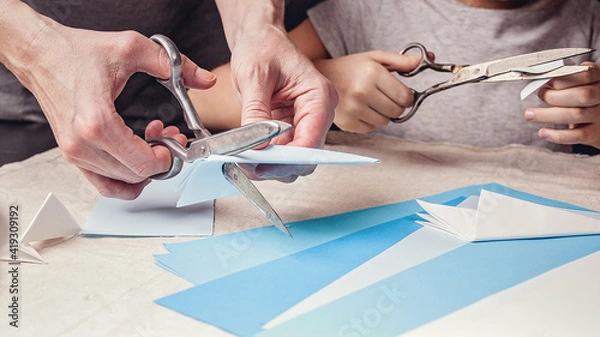 Obraz Hands of a caucasian mother and her daughter are cutting paper snowflakes with scissors. Handmade, christmas decorations, family winter activity. Close-up.