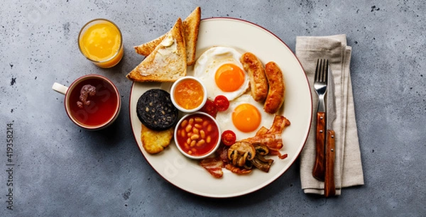 Fototapeta Full fry up English breakfast with fried eggs, sausages, bacon, black pudding, beans, toasts and tea on gray concrete background