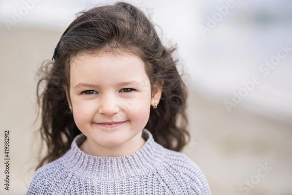 Fototapeta Closeup view portrait of cute calm pretty white smiling happy little girl with long brown hair standing outdoor on sunny spring sandy beach. Sweet child looking in camera happily